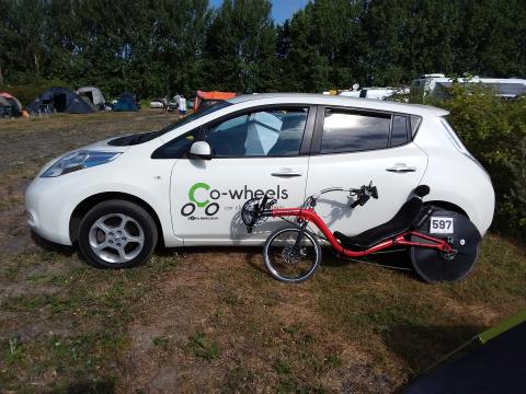 Optima Baron recumbent bicycle with race number 597 leaning against a white Nissan Leaf with a Co-Wheels Birmingham logo on the door panel