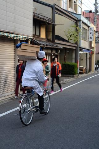 A Japanese man cycling along while holding 2 trays of food he is delivering.