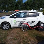 Optima Baron recumbent bicycle with race number 597 leaning against a white Nissan Leaf with a Co-Wheels Birmingham logo on the door panel